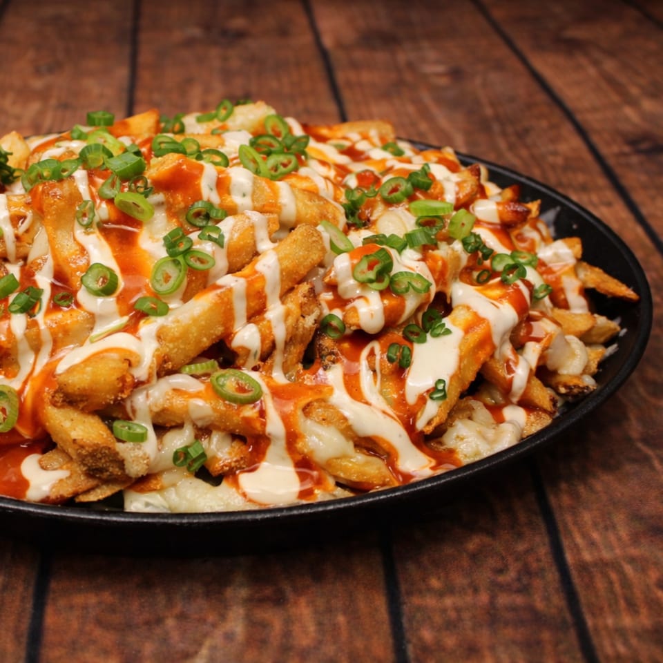 A black plate filled with golden french fries, drizzled with white and orange sauces, and topped with green onions.